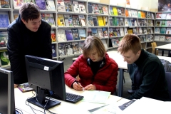 Dwóch licealistów siedzi przy stole z monitorem, klawiaturą i kartkami. Jeden trzyma długopis. Trzeci pochyla się nad nimi. W tle regały z czasopismami.
