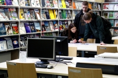 Dwóch chłopców i jedna dziewczyna pochylają się nad stołem, na którym leżą kartki i okulary. W tle widać regały z czasopismami i stoły z monitorami.