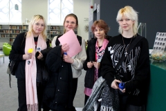 Cztery dziewczyny stoją przy zielonej ladzie. Jedna trzyma różową kartkę, druga magnes z logiem biblioteki i brelok z żółtym kaskiem.