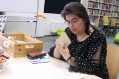 Kobieta w okularach i czarnej bluzce w białe kropki siedzi przy stole i w skupieniu haftuje wzór na kartoniku, tworząc ozdobną kartkę