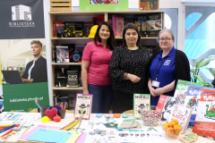 Trzy uśmiechnięte kobiety ze stoiska biblioteki stoją razem przed regałem z książkami