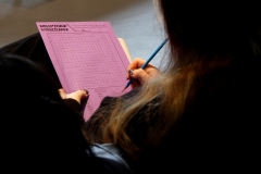 Osoba pokazana od tyłu rozwiązuje fioletową biblioteczną wykreślankę słowną, trzymając w dłoni niebieski ołówek