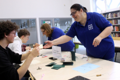 Osoba prowadząca podaje materiał uczestnikom przy stole z nićmi i kartonikami