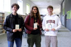 Trzech uśmiechniętych chłopaków stoi w bibliotece, prezentując wykonane przez siebie kartki z haftowanymi motywami kwiatowymi