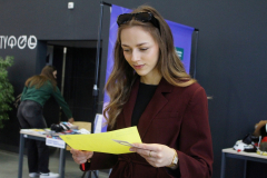 Młoda kobieta w bordowej marynarce i okularach przeciwsłonecznych na głowie uśmiecha się, czytając żółtą kartkę