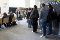 Grupa młodzieży stoi w bibliotece, słuchając uśmiechniętej pracowniczki wypowiadającej się na tle tablicy informacyjnej i regałów z książkami