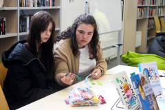 Dwie dziewczyny siedzą przy stole i rozmawiają ze sobą, trzymając w dłoniach przygotowane do haftu kolorowe nici