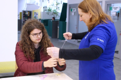 Chłopak w długich kręconych włosach skupia się na szyciu, a pracowniczka w niebieskiej koszulce z logo biblioteki pomaga mu odmierzyć odpowiednią długość nitki