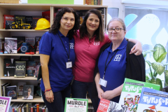 Trzy uśmiechnięte pracowniczki biblioteki stoją obok siebie na stoisku promocyjnym z książkami