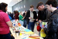 Młodzież przegląda ulotki stojąc i rozmawiając przy stoisku promującym bibliotekę z pracowniczką ubraną w różową koszulkę