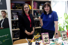 Dwie uśmiechnięte kobiety ze stoiska biblioteki stoją na tle regału z książkami, jedna w czarnej marynarce, druga w granatowej koszulce