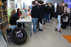 Uczniowie zgromadzeni obok pracowniczki przy stoisku, przed którym na podłodze umieszczono prawdziwą czarną oponę samochodową