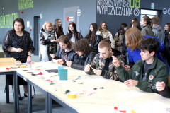 Uczestnicy przy stole wyszywają kartki, w tle grupa obserwatorów