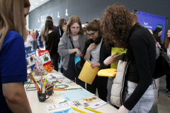 Osoby oglądają materiały na stoisku biblioteki; na stole ulotki, kredki i drobne gadżety