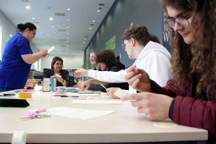 Uczestnicy przy stole wyszywają kartki, prowadząca tłumaczy