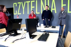 Trzech mężczyzn stojących pod ścianą. Z lewej – mówiąca do nich bibliotekarka. Na pierwszym planie stanowiska komputerowe. 