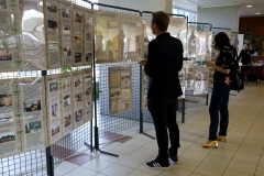 Z lewej strony rząd ekspozytorów. Wiszą na nich antyramy , a w nich wystawa zdjęć historycznych o bibliotece z podpisami. Wystawę oglądają dwie osoby.