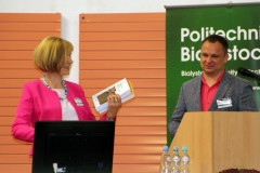Dyrektor Biblioteki Maria Czyżewska trzyma książki i uśmiecha się do mężczyzny, który stoi za mównicą.