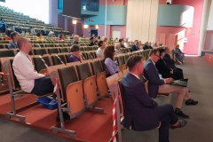 Na auli siedzi w odstępach około 50 osób. Ludzie są zwróceni w kierunku mównicy.