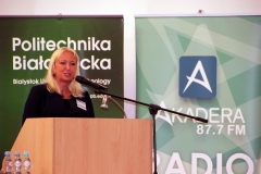 Aula. Przy mównicy stoi kobieta o blond włosach. Przemawia do zebranych. Za nią dwa zielone roll-upy.