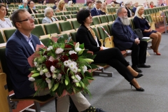 Aula wypełniona siedzącymi w odstępach ludźmi. Na pierwszym planie mężczyzna w ciemnym garniturze z dużym bukietem kwiatów.