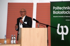 Aula. Za mównicą stoi mężczyzna w ciemnej marynarce i białej koszuli. Za nim stoi zielony roll-up z napisem Politechnika Białostocka.