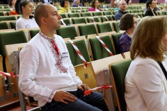 Młody mężczyzna siedzący w białej koszuli z folkowym haftem w kolorze czerwonym. Wokół niego w rzędach siedzą zasłuchani ludzie.