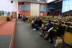 Pomieszczenie auli. Z prawej strony rzędy krzeseł, na których siedzą uczestnicy konferencji. Z lewej strony długa katedra.