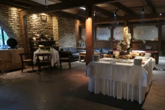 Sala restauracyjna ze stołami przykrytymi białymi obrusami, zastawionymi talerzami z jedzeniem.  Na środku drewniany słup podtrzymujący strop.