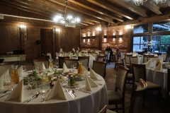 Sala restauracyjna ze stołami przykrytymi białymi obrusami, zastawionymi talerzami z jedzeniem. Widoczne drewniane belki stropowe i żyrandol.