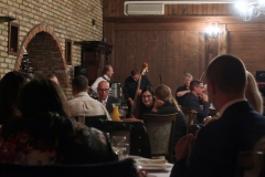 Sala restauracyjna. Na pierwszym planie dużo osób siedzących przy stołach, rozmawiających. W tle 3-osobowy zespół muzyczny, kobieta i dwóch mężczyzn.