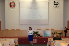 Aula z rzędami krzeseł i długą katedrą. Przed nią stoi kobieta w białej bluzce i granatowej spódnicy z mikrofonem w prawej ręce i kartką w lewej.