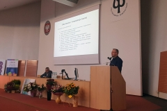 Na pierwszym planie mównica, z której przemawia mężczyzna. Do niej przylega katedra, za którą siedzi drugi mężczyzna. Nad katedrą ekran projekcyjny.