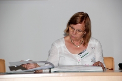 Kobieta w okularach o brązowych włosach do ramion siedzi za biurkiem i przegląda dużą księgę. Ma pochyloną w dół głowę. W prawej ręce trzyma długopis.