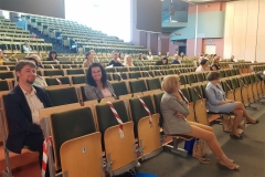Aula z rzędami krzeseł, na których siedzą osoby, w tym uśmiechnięta kobieta z długimi, kręconymi, ciemnymi włosami oraz uśmiechnięty mężczyzna.