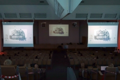 Aula. W połowie sali widać ekrany po obu stronach, na środku duży ekran główny. Na ekranach wyświetlany jest obraz kamienicy.