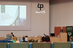 Aula. Dwie kobiety zwrócone przodem do słuchaczy. Kobieta po prawej ma ciemne włosy i czarno-białą bluzkę. Druga siedzi ze spuszczoną głową.