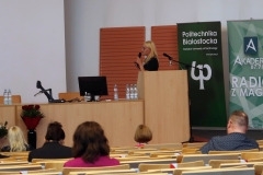 Aula. Przy mównicy stoi kobieta o blond włosach i w czarnej sukience. Za nią dwa zielone roll-up - Akadera 87.7 FM i Politechnika Białostocka.