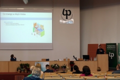 Aula. Mężczyzna zwrócony przodem do słuchaczy stoi przy mównicy i w ręku trzyma mikrofon. Kobieta obok niego jest odwrócona w kierunku ekranu.