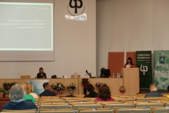 Aula. Kobieta po prawej mówi do mikrofonu umieszczonego na mównicy. Ma ciemne włosy i czerwoną marynarkę. Druga kobieta siedzi ze spuszczoną głową.