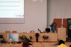 Pomieszczenie auli  z rzędami krzeseł, na których siedzą osoby. Na środku katedra. Za nią stoi mężczyzna z mikrofonem. Nad katedrą ekran projekcyjny.
