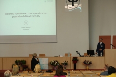 Aula z rzędami krzeseł, na których siedzą osoby. Na środku katedra. Za nią stoi mężczyzna, przed nią kobieta z mikrofonem. Na ścianie ekran projekcyjny.