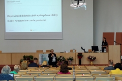 Aula z rzędami krzeseł, na których siedzą osoby. Na środku długa katedra, za którą stoi kobieta, przed nią kobieta z mikrofonem.