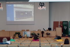 Aula z rzędami krzeseł, na których siedzą osoby. Na środku katedra, nad którą wisi ekran projekcyjny. Z lewej strony stoi kobieta.