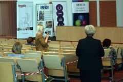 Aula z rzędami krzeseł, na których siedzą osoby. Z Lewej strony widać kobietę z mikrofonem, z prawej kobietę stojącą tyłem.
