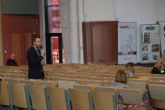 Aula z rzędami krzeseł, na których siedzą osoby. Z Lewej strony widać stojącego bokiem mężczyznę w czarnej marynarce i mikrofonem w ręku.