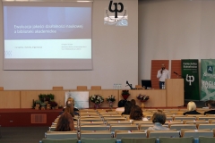 Aula z krzesłami, na których siedzą osoby. Na środku katedra. Za nią stoi mężczyzna w białej koszuli z mikrofonem w ręce. Na ścianie ekran projekcyjny.
