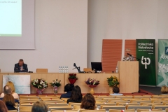 Aula z rzędami krzeseł, na których siedzą osoby. Na środku katedra, za którą siedzi  mężczyzna. Z mównicy, przylegającej do katedry, przemawia kobieta.