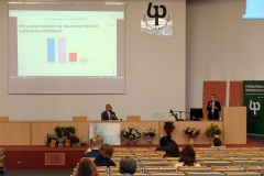 Aula z rzędami krzeseł, na których siedzą osoby. Na środku katedra, za którą siedzi  mężczyzna. Z mównicy, przylegającej do katedry, przemawia kobieta.