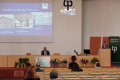 Aula z rzędami krzeseł, na których siedzą osoby. Na środku długa katedra, za którą siedzi mężczyzna. Drugi przemawia z mównicy, przylegającej do niej.